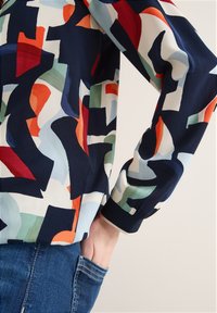 Personne portant une chemise à manches longues avec un motif abstrait multicolore, la main rentrée dans la poche d'un jean en denim bleu.