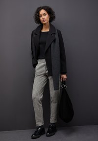 Czarny wełniany płaszcz z szarym szalikiem, czarna bluzka, szare w kratkę spodnie i czarne buty. Modelka trzyma czarną torebkę.