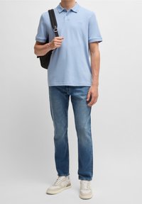 Světlomodrá polokošile s límečkem, černá výšivka, v kombinaci s modrými džínami a krémovými teniskami. Křížová taška visí přes rameno.