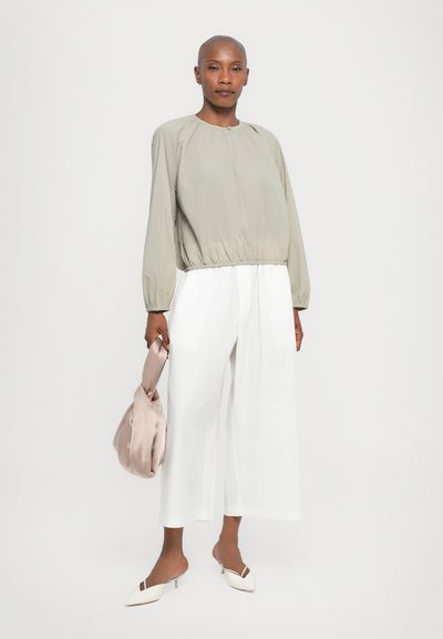 Blusa verde chiaro a maniche lunghe con orlo arricciato, abbinata a pantaloni bianchi a gamba larga. Il modello tiene una borsa rosa chiaro e indossa mule bianchi.