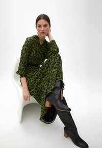 Vihreä leopardikuosiset mekko, joka on valmistettu kevyestä kankaasta ja jossa on pitkät hihat, yhdessä polven ylle ulottuvien mustien saappaiden kanssa, joissa on kasattu korko.