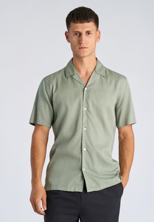 Koszula z krótkim rękawem w jasnozielonym kolorze, z kołnierzem typu camp, o gładkiej fakturze i białych guzikach. Noszona z czarnymi spodniami, widok z przodu.
