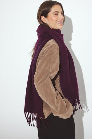 Sciarpa di lana viola con dettagli frangiati drappata su una giacca in suede beige, indossata da un modello su uno sfondo neutro.