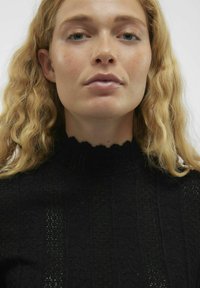 Zwarte textuurtrui met ribbelontwerp, gegolfde halslijn en opengewerkte patroon. Model poseert met lichtblond, golvend haar.