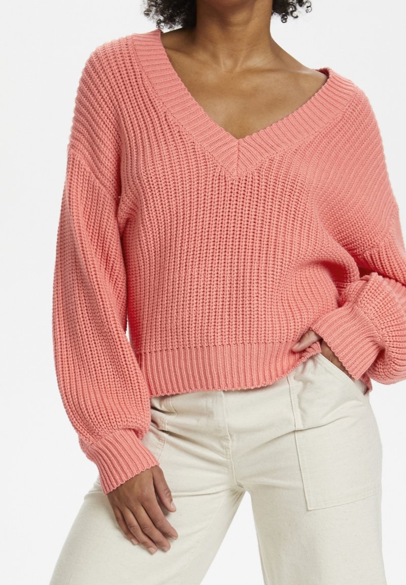 Maglione rosa a maglia lavorata con scollo a V e maniche a palloncino, abbinato a pantaloni chiari. Presenta polsini e orlo a coste.