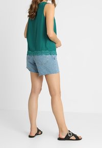 Top verde senza maniche con bordo in pizzo, abbinato a pantaloncini di jeans azzurri chiari. Sandali neri con dettaglio circolare in metallo.