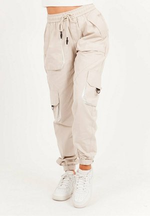 Beige cargobroek met elastische tailleband, meerdere zakken, reflecterende accenten en taps toelopende boorden, gecombineerd met witte sneakers.