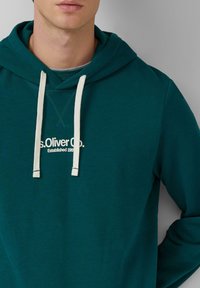 Mees, kellel on seljas tealds sinine kapuutsiga sweatshirt, millel on valged nöörid ja rinnas on valgetes tähtedes kirjutatud "s.Oliver Co. Established 1969".