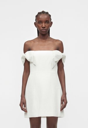 Fiatal nő fonott hajjal, fehér váll nélküli ruhát visel, amely íj díszítéssel van az felső karokon, sima világos háttér előtt.