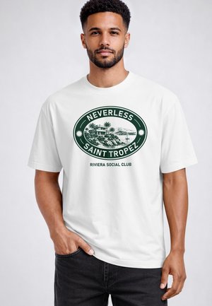 Mann mit kurzen lockigen Haaren und Bart, der ein weißes T-Shirt mit grünem Aufdruck trägt, das ein Küstenhaus, Palmen, ein Segelboot und den Text "Nevertheless Saint Tropez Riviera Social Club" zeigt.