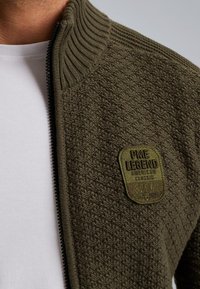 Giacca zip-up verde oliva con tessuto texturizzato, colletto a costine e una toppa con "PME LEGEND AMERICAN CLASSIC 52941" ricamata.