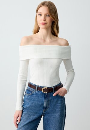 Top bianco a costine senza spalle con maniche lunghe, abbinato a jeans blu a vita alta e una cintura marrone con fibbia dorata.