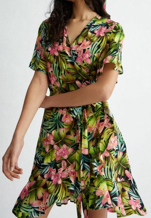 Vrouw draagt een korte mouw wikkeljurk met tropische roze bloemen en groene bladeren, gestrikt in de taille, met gerimpelde zoom.