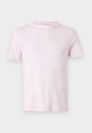 Mladý muž má na sebe svetloružové tričko, krémové kraťasy po kolená a biele slip-on topánky, stojí s rukami v vreckách.