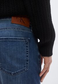 Σκούτερο πλεκτό πουλόβερ, μπλε τζιν με δερμάτινη ετικέτα Calvin Klein· ορατή τσέπη με καθορισμένο ραφή και ξεθωριασμένη υφή.