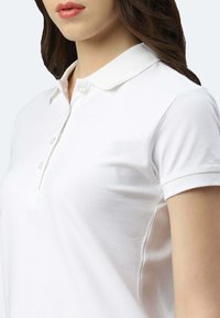 Frau trägt ein weißes Poloshirt mit kurzen Ärmeln, einem Kragen und drei Knöpfen, dargestellt von der Schulter bis zum Kinn.