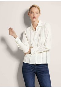 Witte overhemd met ruches en geborduurde accenten aan de zijkanten, gecombineerd met donkerblauwe slim fit spijkerbroek.