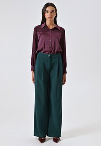 Blusa di raso viola con design a bottoni, abbinata a pantaloni a gamba larga verdi a vita alta con un passante rotondo per la cintura.