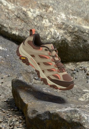 Кафяво и оранжево туристическо обувка с подметка Vibram, плаваща над скалист терен.