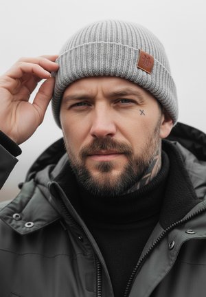 Gray knit beanie with a ribbed texture and brown leather patch. Worn by a person in a black jacket and sweater, with a neutral background.