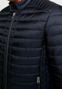 Marinblå puffjacka med horisontell sömnad, hög krage, dragkedjeöppning framtill och sidofickor med silverfärgade detaljer.