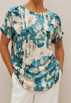 Kobieta ubrana w krótki rękaw z abstrakcyjnym kwiatowym wzorem w kolorach teal, beżowym i białym, z kieszenią na piersi oraz białe spodnie.