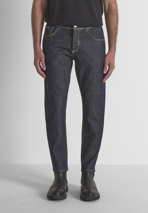 Man draagt donkerblauwe rechte jeans, zwarte laarzen en een zwart overhemd, staand tegen een effen lichte achtergrond.