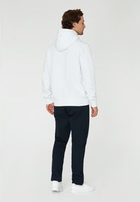 Sudadera blanca con capucha de textura suave, puños y dobladillo de tipo acanalado; combinada con pantalones oscuros y zapatillas blancas. Vista trasera.