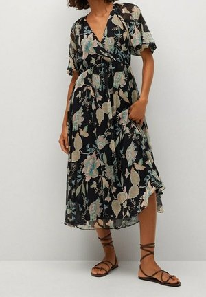 Frau trägt ein schwarzes Midikleid mit großem pastellfarbenem Blumenmuster, V-Ausschnitt, kurzen Ärmeln und braunen flachen Schnürsandalen, stehend vor einer weißen Wand.