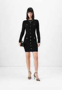 Donna che indossa un vestito cardigan aderente nero con bottoni dorati, sandali neri con tacco alto, collana a catena dorata e tiene una borsa nera.