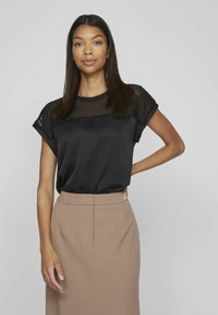 Blusa nera con un pannello superiore trasparente, maniche corte arrotolate, abbinata a una gonna a tubino marrone chiaro. Texture liscia e design semplice.