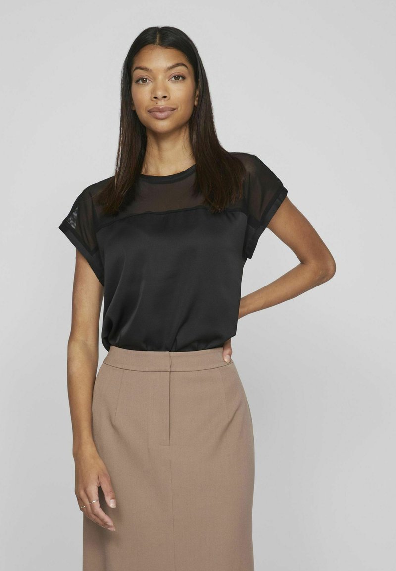 Blusa nera con un pannello superiore trasparente, maniche corte arrotolate, abbinata a una gonna a tubino marrone chiaro. Texture liscia e design semplice.