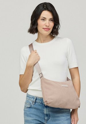 Woman with short dark hair wearing a white short-sleeve top and blue jeans, holding a light pink corduroy crossbody bag over her shoulder.
