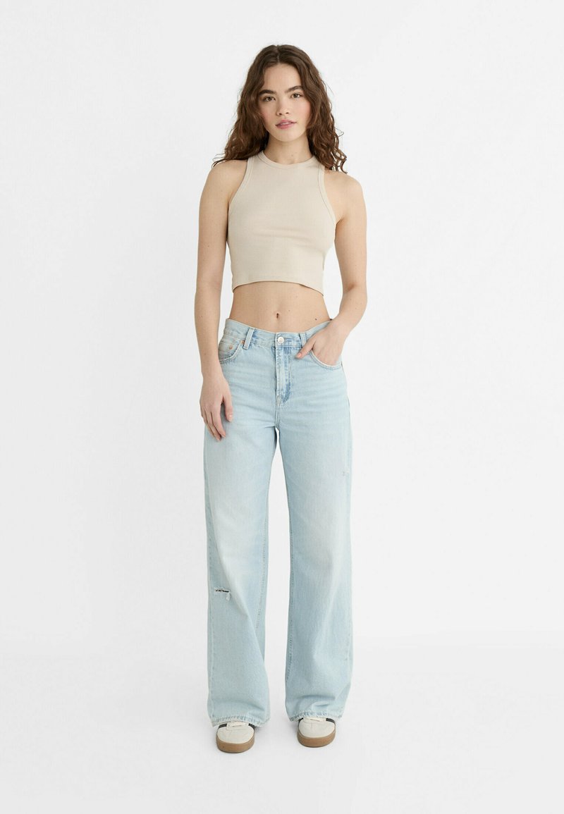 Top tank beige accorciato abbinato a jeans a gamba larga di un blu chiaro. I jeans presentano dettagli scoloriti e un design a vita alta. Le sneakers bianche completano l'outfit.