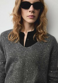 Maglione grigio scintillante con scollo a V e maniche lunghe, realizzato in un materiale testurizzato, indossato sopra una camicia nera con colletto.