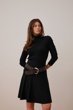 Frau trägt ein schwarzes langärmeliges Kleid mit breitem braunem Gürtel, steht mit den Händen hinter dem Rücken und schaut seitlich vor einem einfarbigen Hintergrund.