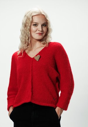 Kobieta o falowanych blond włosach, ubrana w jasnoczerwony, dziergany sweter z złotą broszką w kształcie serca, z rękami w kieszeniach czarnych spodni, na jednolitym tle.