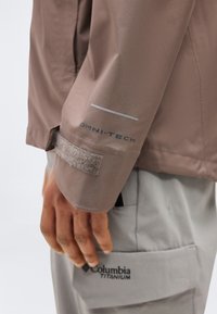 Manche de veste marron avec le texte "OMNI-TECH" et une sangle à velcro, reposant au-dessus d'une poche de pantalon gris étiquetée "Columbia Titanium".