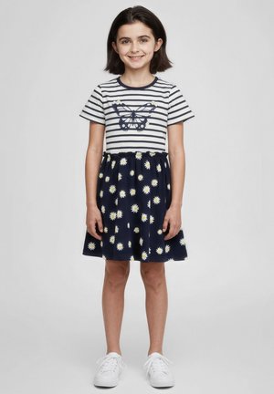 Junges Mädchen lächelt, trägt ein marineblau-weiß gestreiftes Oberteil mit Schmetterling, einen marineblauen Rock mit weißen Gänseblümchen und weiße Turnschuhe, steht vor grauem Hintergrund.