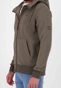 Felpa verde oliva con zip, cappuccio con cordino, tasche a canguro, polsini a coste e un piccolo logo sulla manica. Materiale in misto cotone.