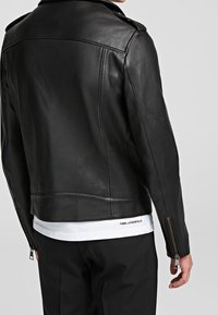 Svart läderjacka med en figurpassad design, slät yta, dragkedjeärmar och en skräddarsydd silhuett. Vit skjorta med logotyp synlig nedanför.