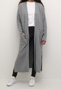 Long, gray ribbed cardigan with open front, side slits, and two front pockets, paired with black pants and white sneakers.