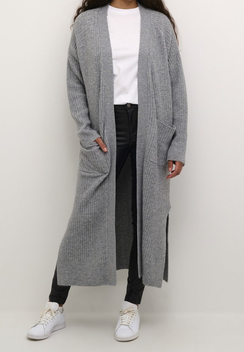 Long, gray ribbed cardigan with open front, side slits, and two front pockets, paired with black pants and white sneakers.