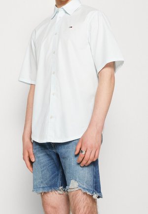 Homme portant une chemise blanche à manches courtes boutonnée et un short en jean effiloché, avec une bague en argent à la main droite.