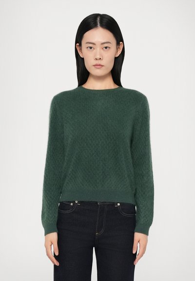 Pull en maille verte avec un motif texturé, col rond et manches longues, associé à un jean foncé. Tissu doux et léger.