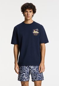 Camiseta de algodón azul marino de manga corta con un diseño gráfico que presenta "Coral Coast" en el frente, combinada con un pantalón corto de baño estampado.