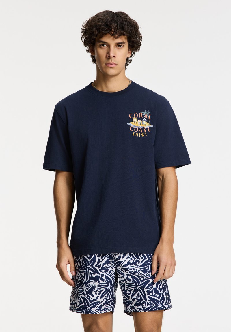 Camiseta de algodón azul marino de manga corta con un diseño gráfico que presenta "Coral Coast" en el frente, combinada con un pantalón corto de baño estampado.