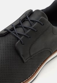 Chaussure en cuir noir avec un design perforé, à lacets plates, et un accent brun sur la semelle. Texture lisse avec des détails de couture soignés.