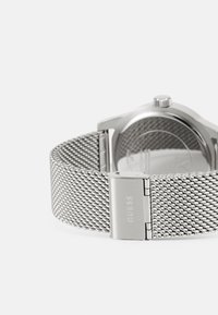 Silbernes Mesh-Uhrarmband mit metallischem Verschluss, ausgestattet mit einem strukturierten Ziffernblatt und einem robusten Gehäuse, das ein schlankes, minimalistisches Design zeigt.