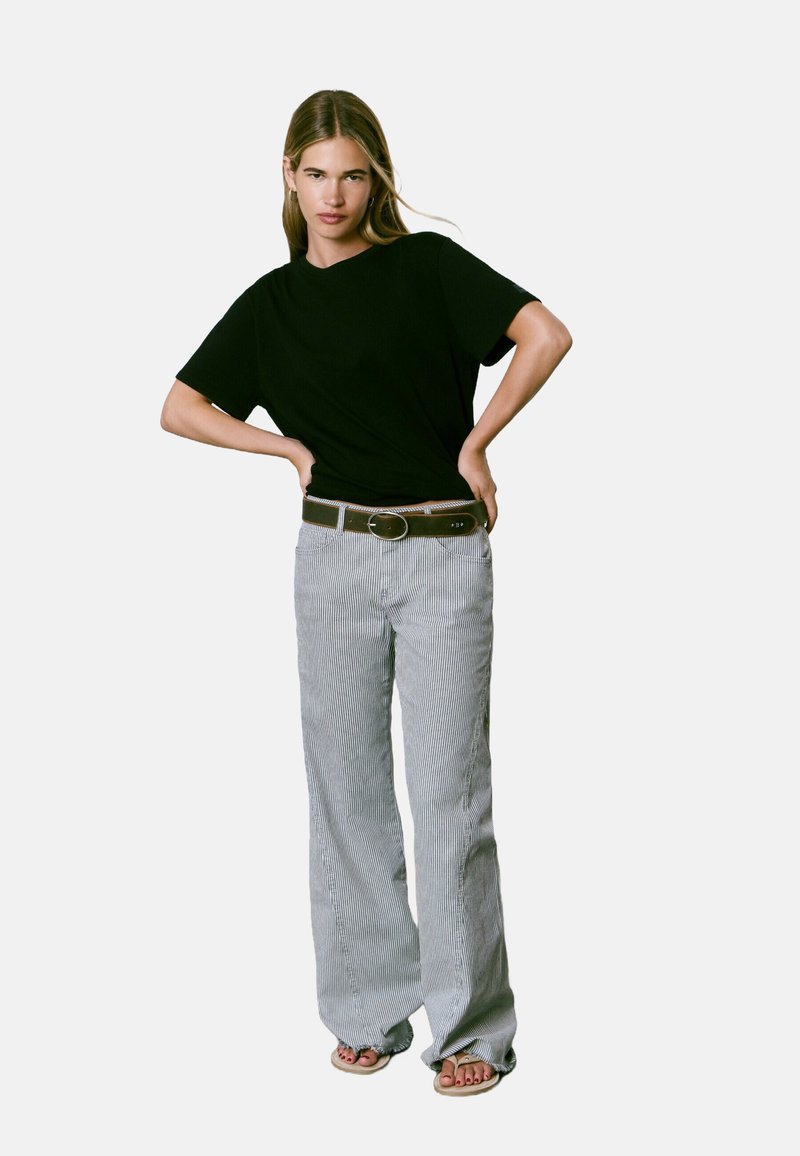 T-shirt noir à manches courtes associé à un pantalon large rayé gris et blanc. La tenue comprend une ceinture marron avec une boucle ronde.